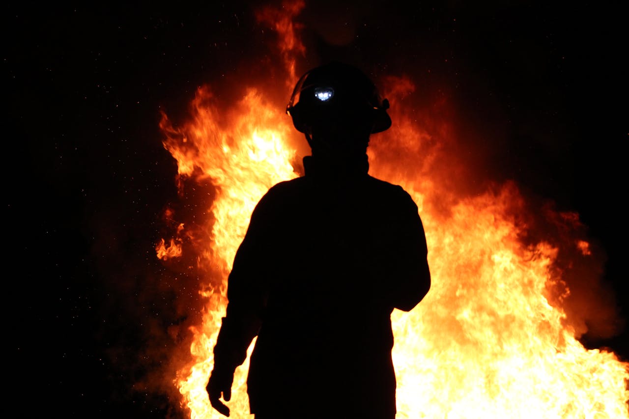 A silhouetted firefighter stands against a backdrop of roaring flames at night.