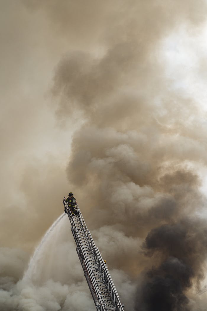 Brave firefighter extinguishing fire from a ladder amidst heavy smoke backdrop.